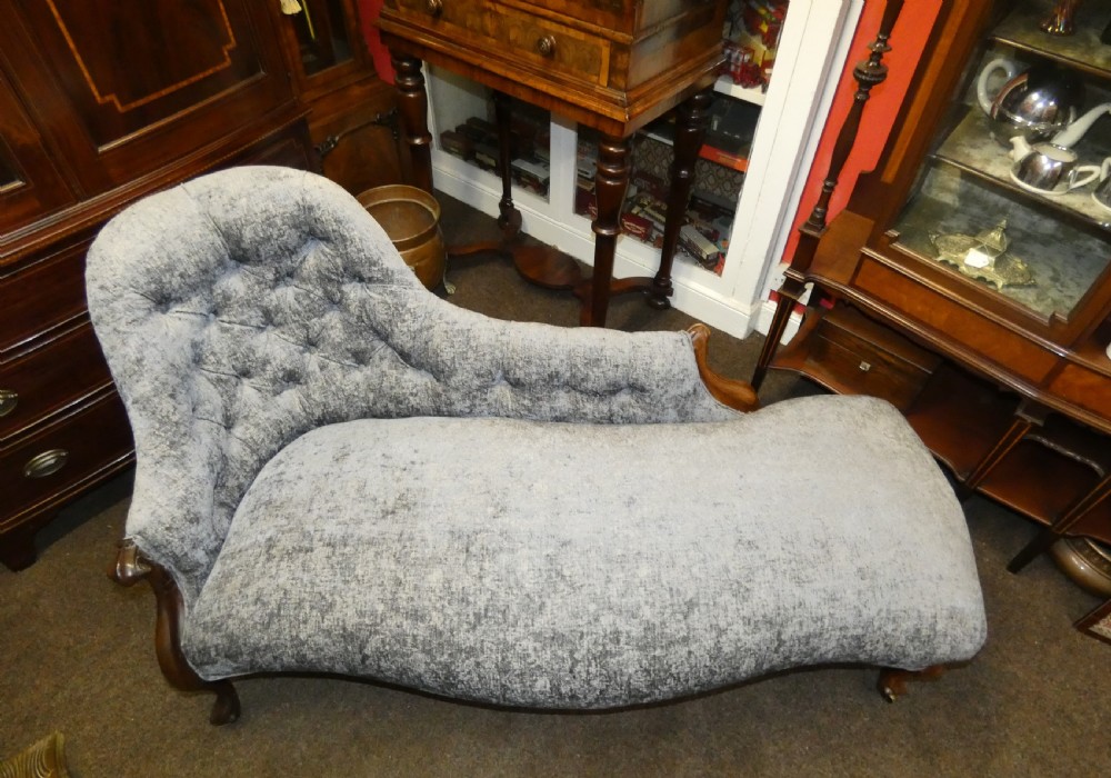A Victorian-style chaise longue upholstered in textured grey fabric featuring a deep button-tufted asymmetrical backrest and carved wooden cabriole legs on brass castors. View VIII