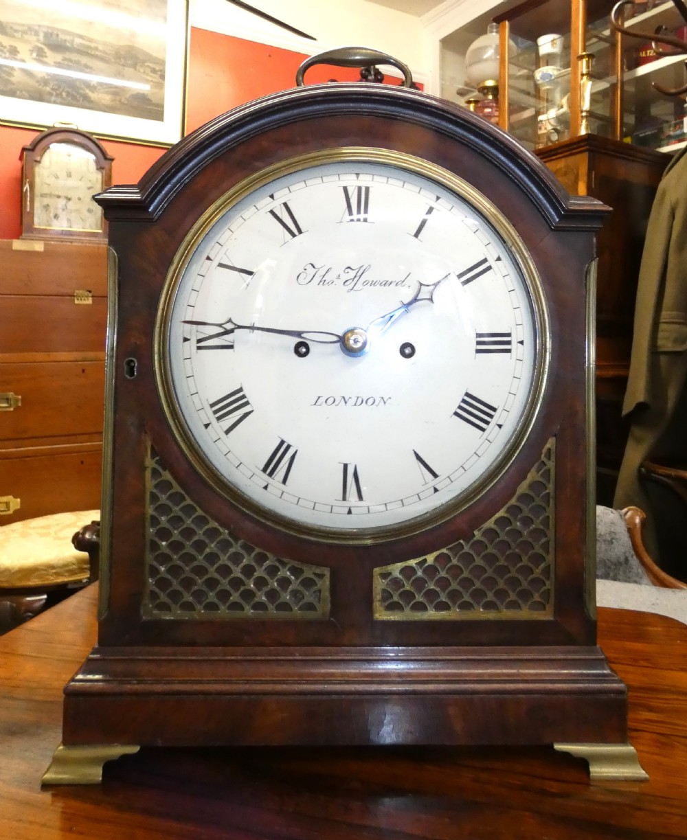 An antique dark mahogany bracket clock featuring an arched top with a brass carrying handle, a circular white dial signed Thos. Howard London, and brass fish-scale grilles.
