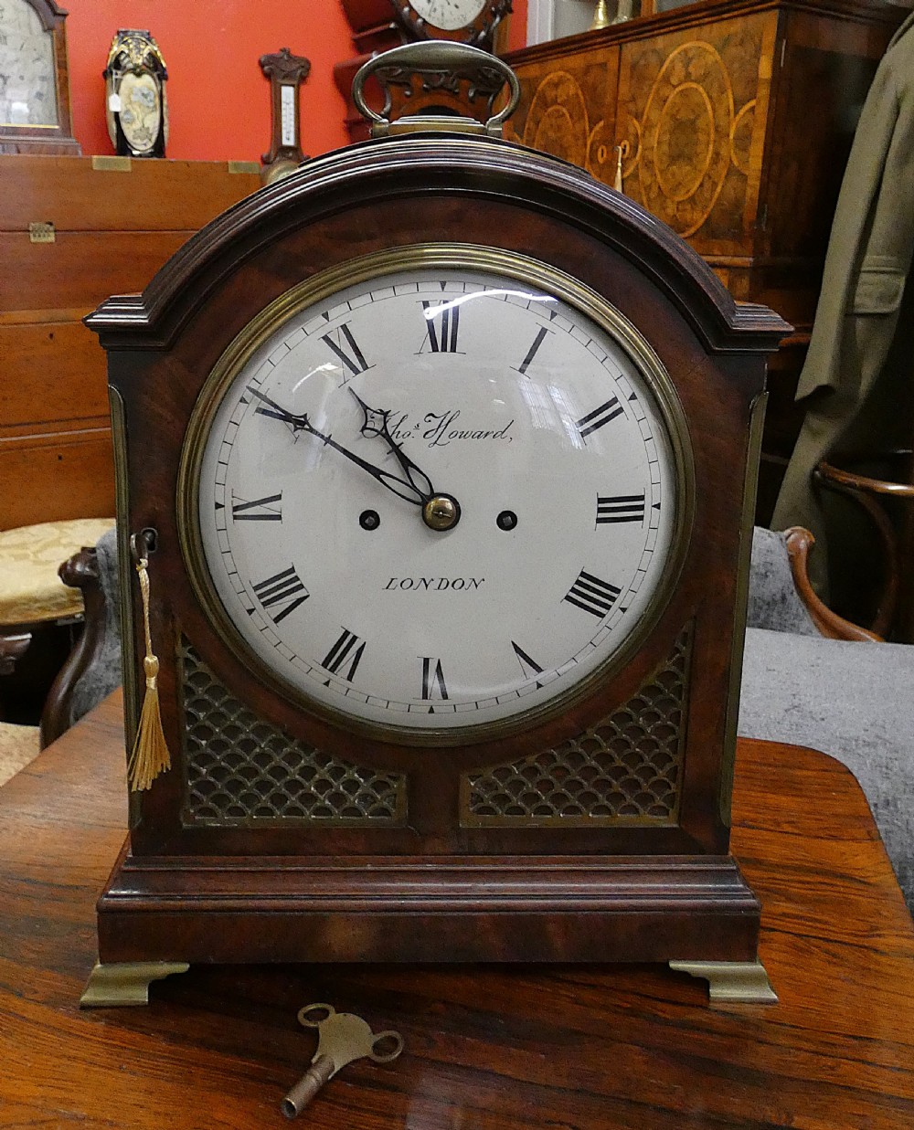 An antique dark mahogany bracket clock featuring an arched top with a brass carrying handle, a circular white dial signed Thos. Howard London, and brass fish-scale grilles. View II