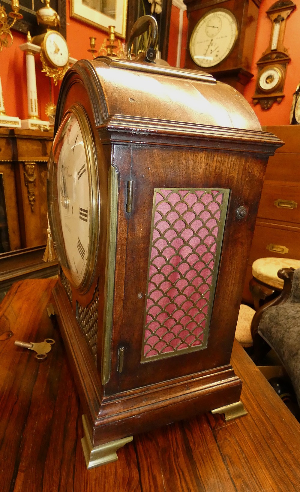 An antique dark mahogany bracket clock featuring an arched top with a brass carrying handle, a circular white dial signed Thos. Howard London, and brass fish-scale grilles. View III