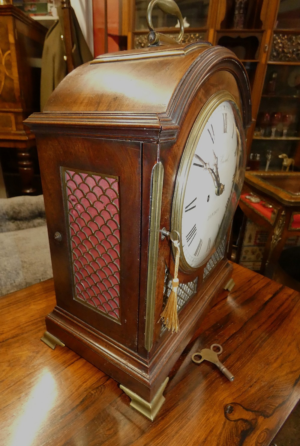 An antique dark mahogany bracket clock featuring an arched top with a brass carrying handle, a circular white dial signed Thos. Howard London, and brass fish-scale grilles. View IV