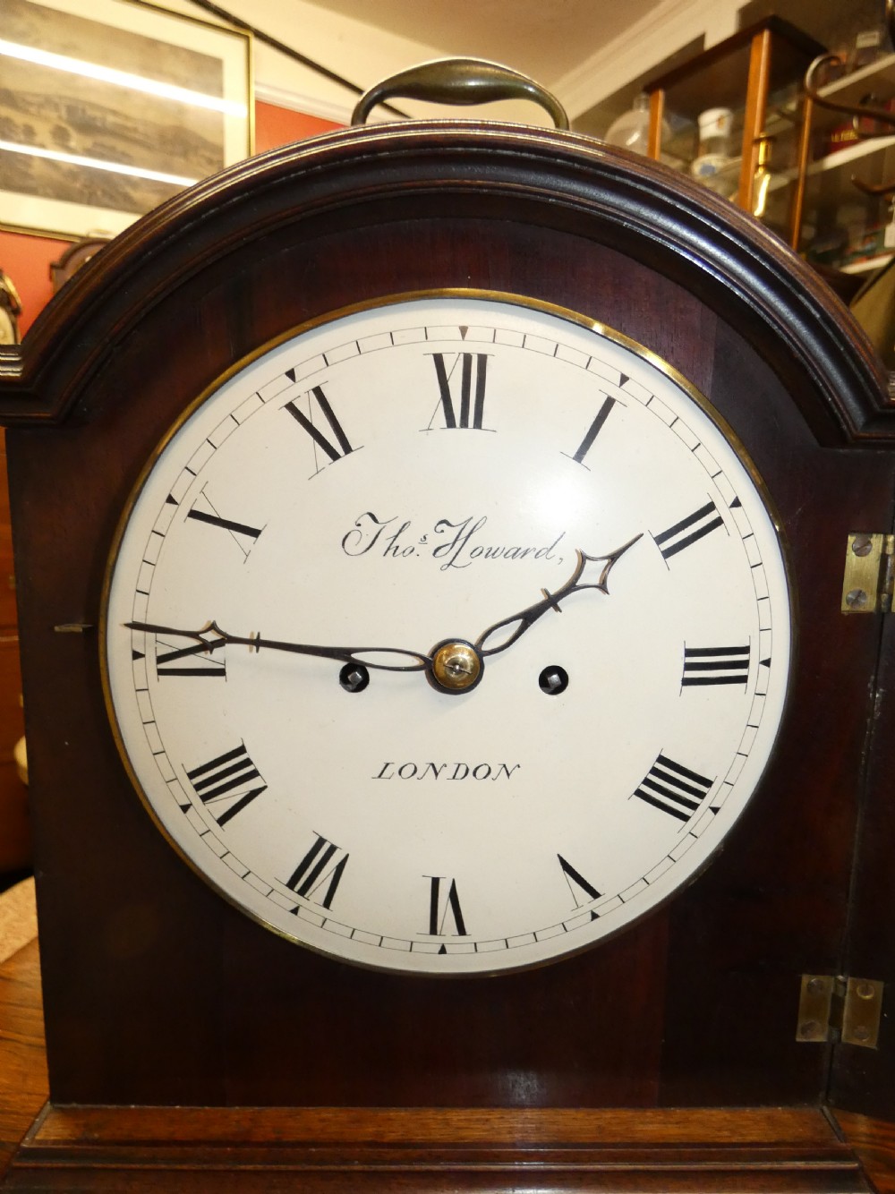 An antique dark mahogany bracket clock featuring an arched top with a brass carrying handle, a circular white dial signed Thos. Howard London, and brass fish-scale grilles. View V