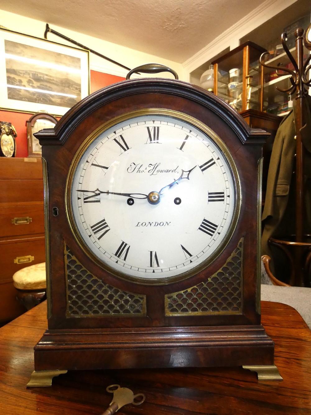 An antique dark mahogany bracket clock featuring an arched top with a brass carrying handle, a circular white dial signed Thos. Howard London, and brass fish-scale grilles. View VI