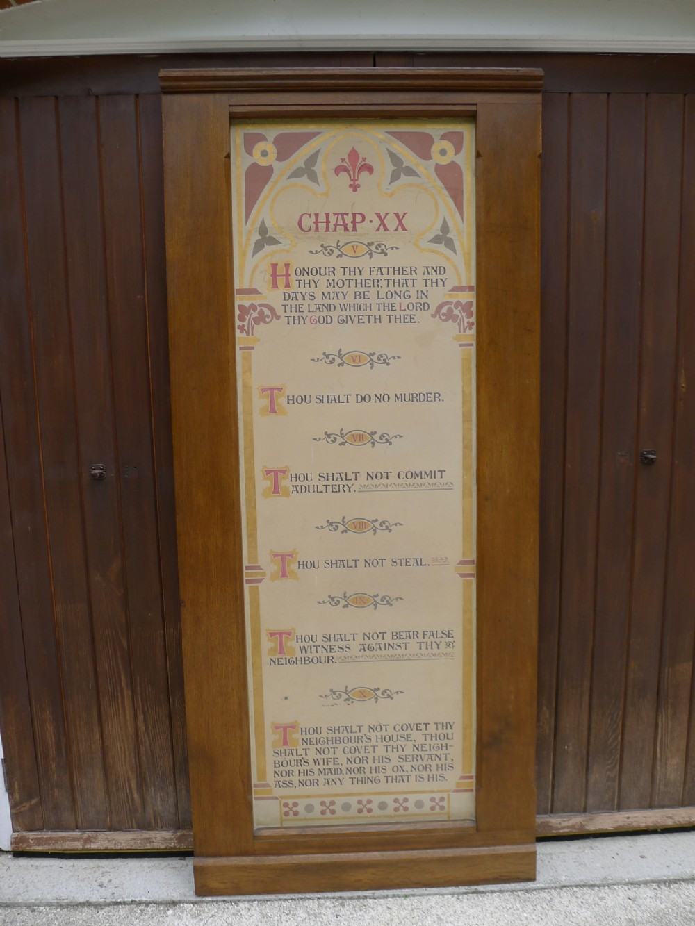 A tall, framed Victorian timber panel displaying painted biblical scriptures with Gothic arches and decorative fleur-de-lis motifs in terracotta and yellow tones.