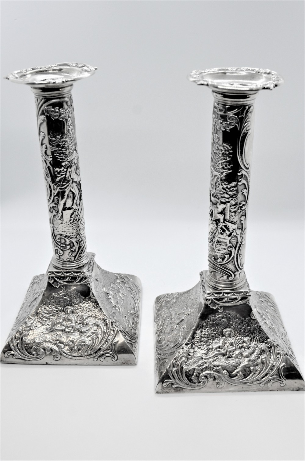 A pair of ornate Victorian silver candlesticks featuring embossed figurative relief scenes along the columns and flared square bases.