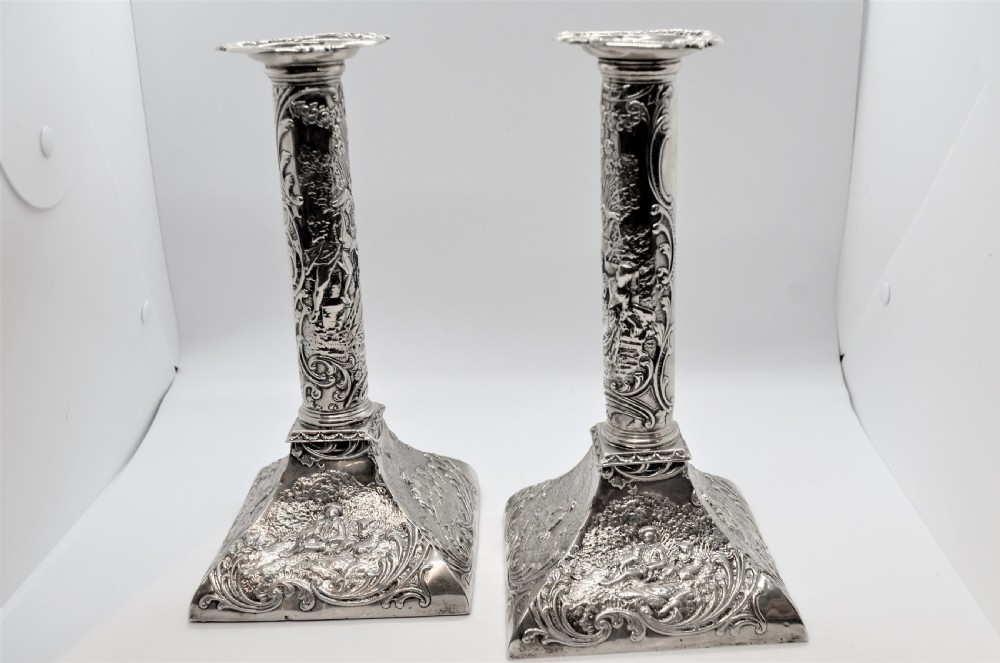 A pair of ornate Victorian silver candlesticks featuring embossed figurative relief scenes along the columns and flared square bases. View II
