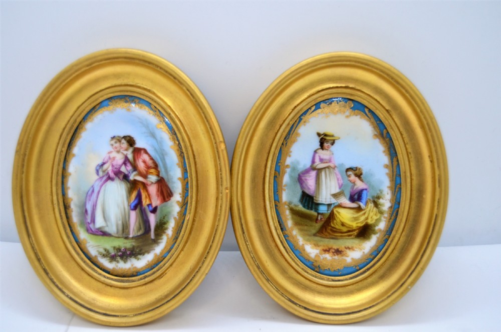 A pair of oval gilt-framed porcelain plaques featuring hand-painted pastoral figures within sky blue and gold scroll borders.