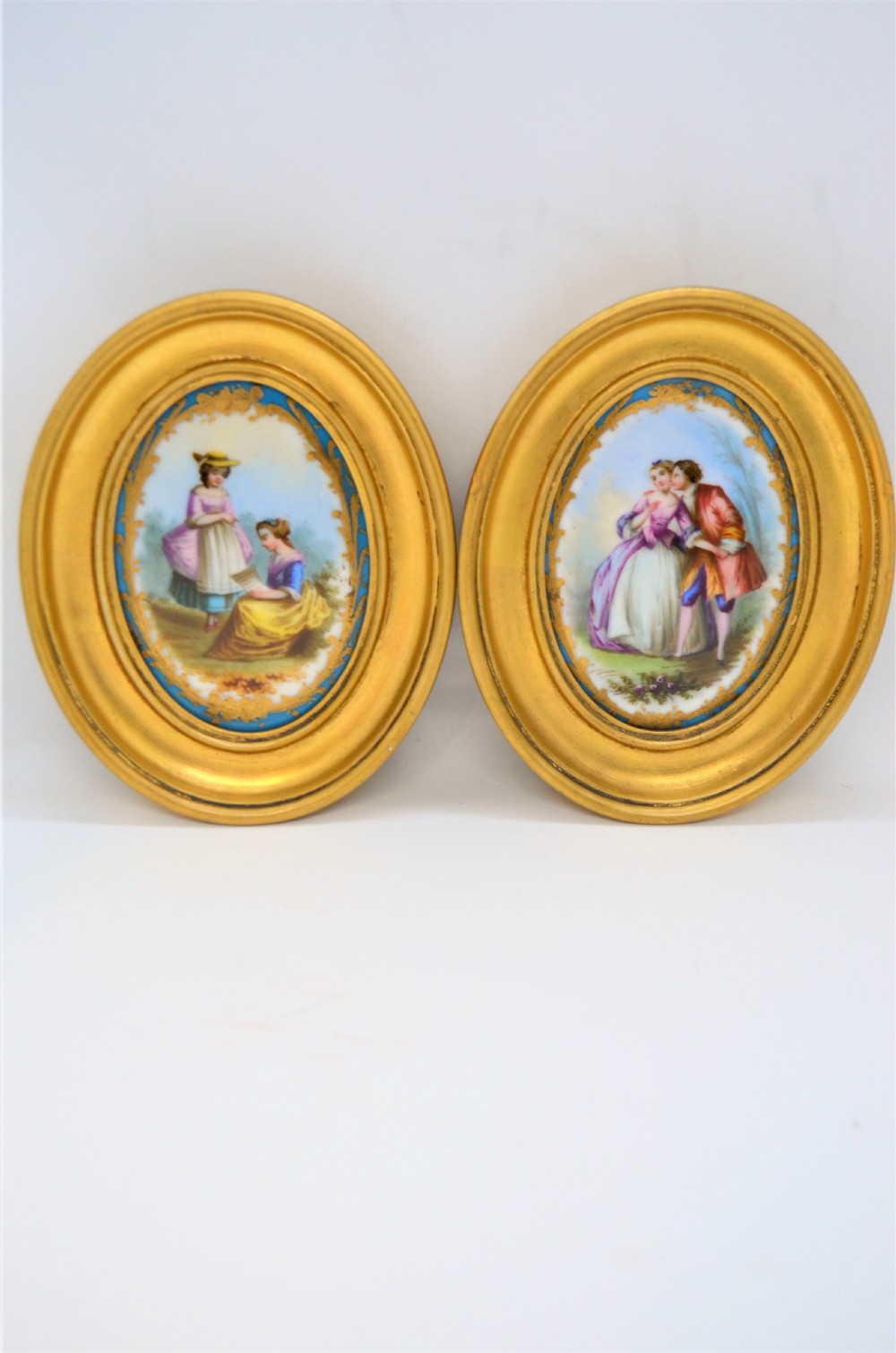 A pair of oval gilt-framed porcelain plaques featuring hand-painted pastoral figures within sky blue and gold scroll borders. View V