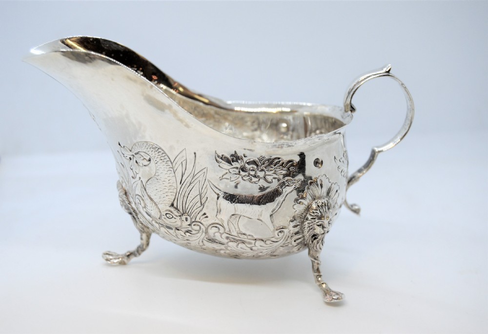 An antique Irish sterling silver cream jug featuring detailed repousse chasing of animals, lion-headed tripod legs, and a scroll handle.