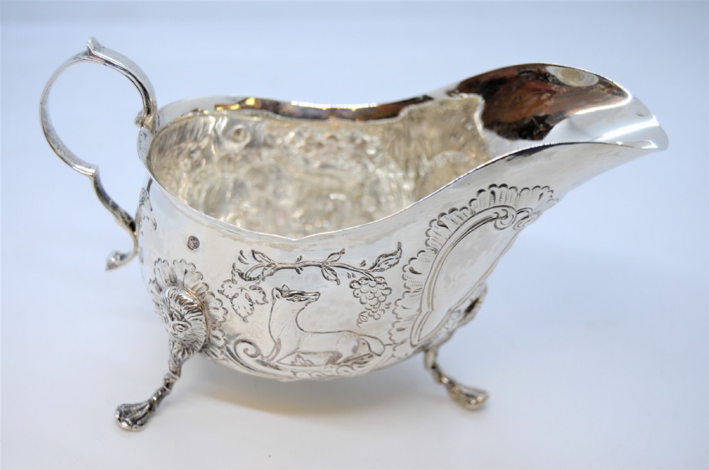 An antique Irish sterling silver cream jug featuring detailed repousse chasing of animals, lion-headed tripod legs, and a scroll handle. View II