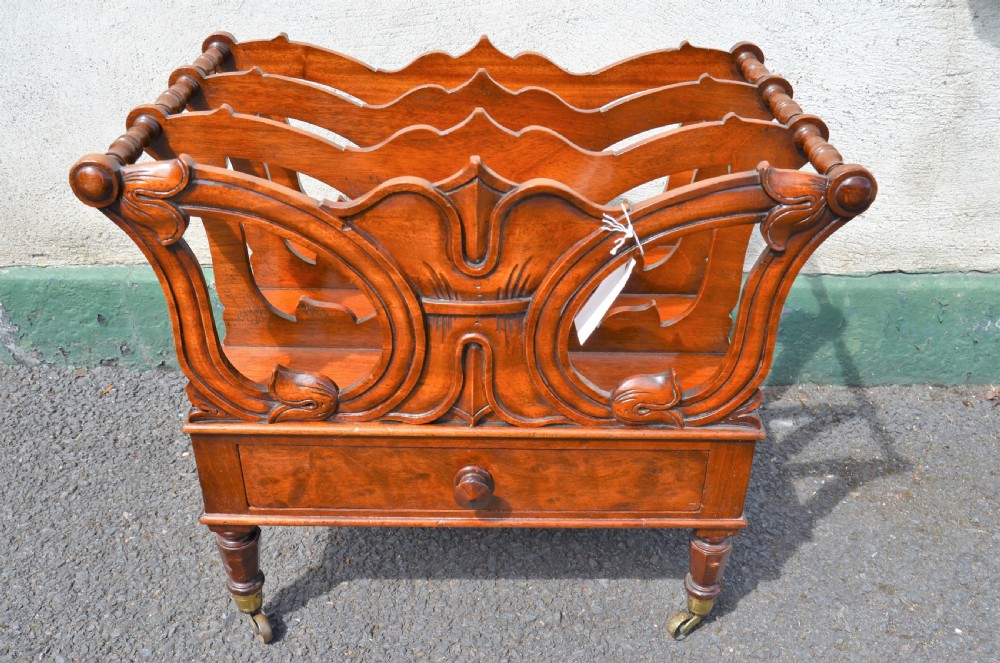 A Victorian mahogany canterbury featuring three carved slotted compartments, a lower drawer with a turned knob, and turned legs with brass castors.