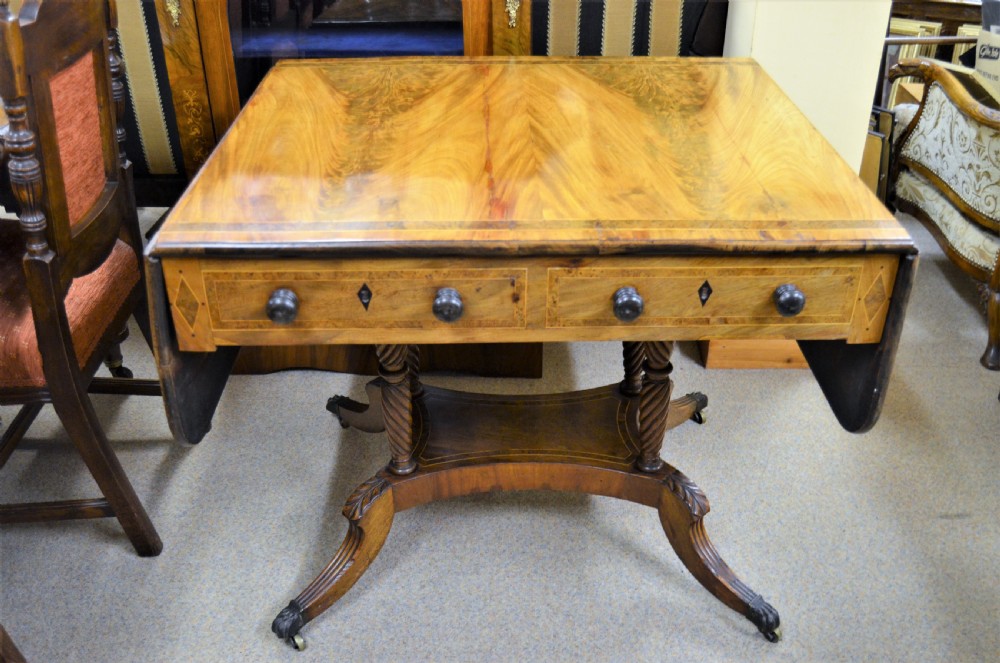 A Regency flame mahogany sofa table featuring two drop-leaf sides, two front drawers with turned wooden knobs, twin spiral-turned pedestal columns, and splayed legs ending in brass castors.