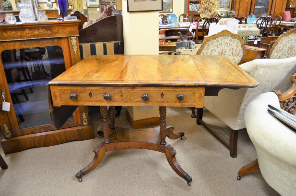A Regency flame mahogany sofa table featuring two drop-leaf sides, two front drawers with turned wooden knobs, twin spiral-turned pedestal columns, and splayed legs ending in brass castors. View II