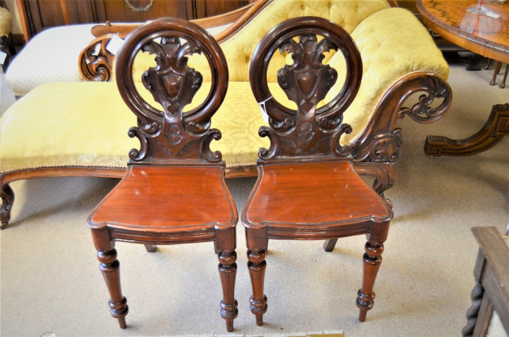 Pair of antique Victorian mahogany hall chairs featuring open carved circular shield backrests, solid timber seats, and turned front legs.