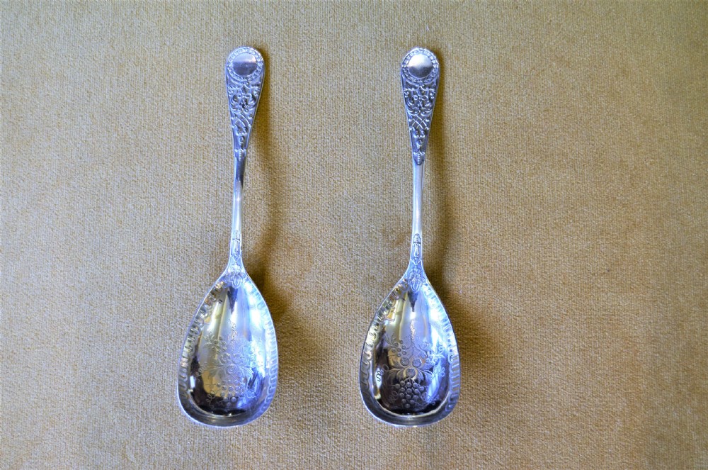 A pair of silver serving spoons featuring detailed fruit engraving in the bowls and decorative handles.