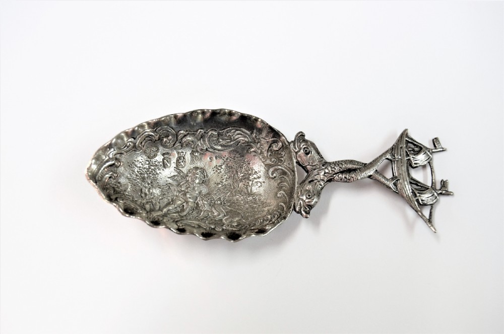 A Dutch silver caddy spoon featuring an embossed figural scene inside the scalloped bowl and a handle formed of entwined sea creatures topped with a sailing ship finial.