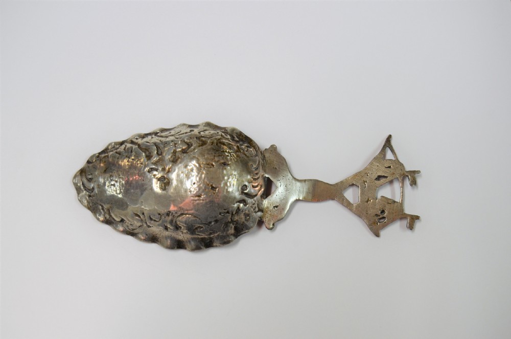 A Dutch silver caddy spoon featuring an embossed figural scene inside the scalloped bowl and a handle formed of entwined sea creatures topped with a sailing ship finial. View II
