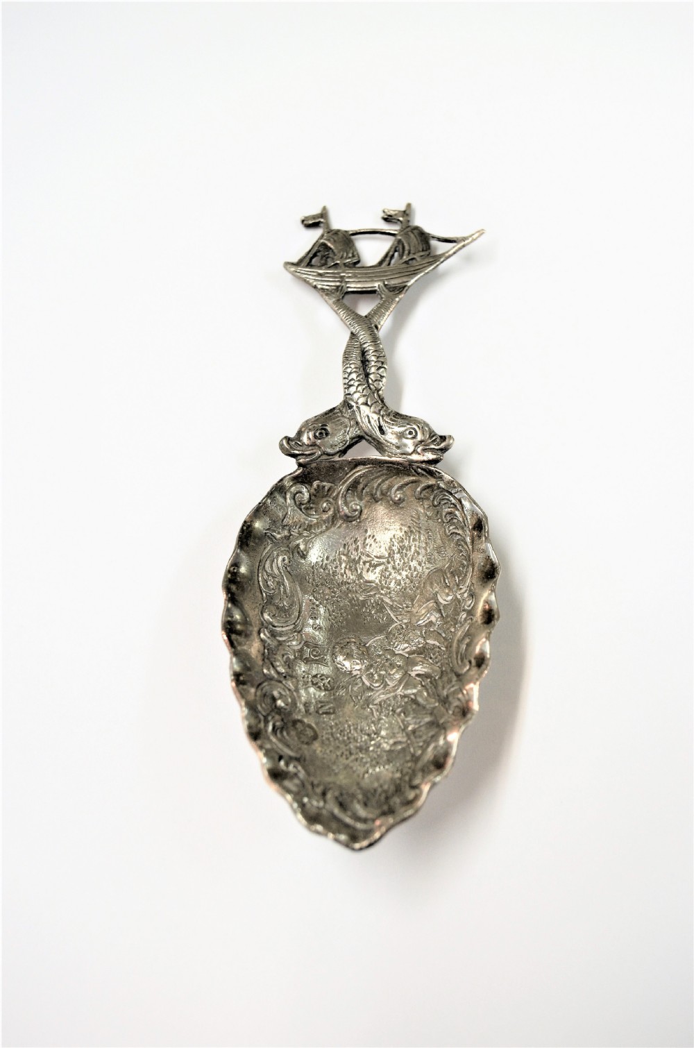A Dutch silver caddy spoon featuring an embossed figural scene inside the scalloped bowl and a handle formed of entwined sea creatures topped with a sailing ship finial. View V