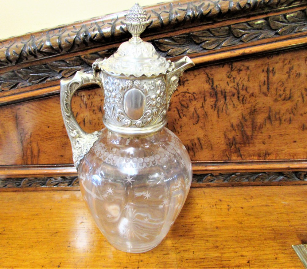 An antique clear glass claret jug decorated with etched star motifs and featuring a silver-plated repousse collar, handle, and finial lid.