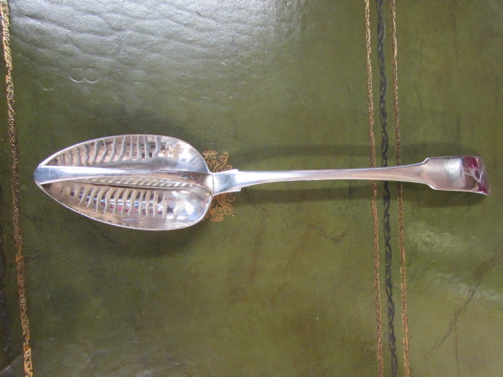 A George III Irish sterling silver strainer spoon featuring a long flat handle with an engraved crest and an oval bowl with a central divider and slotted decorative piercings.