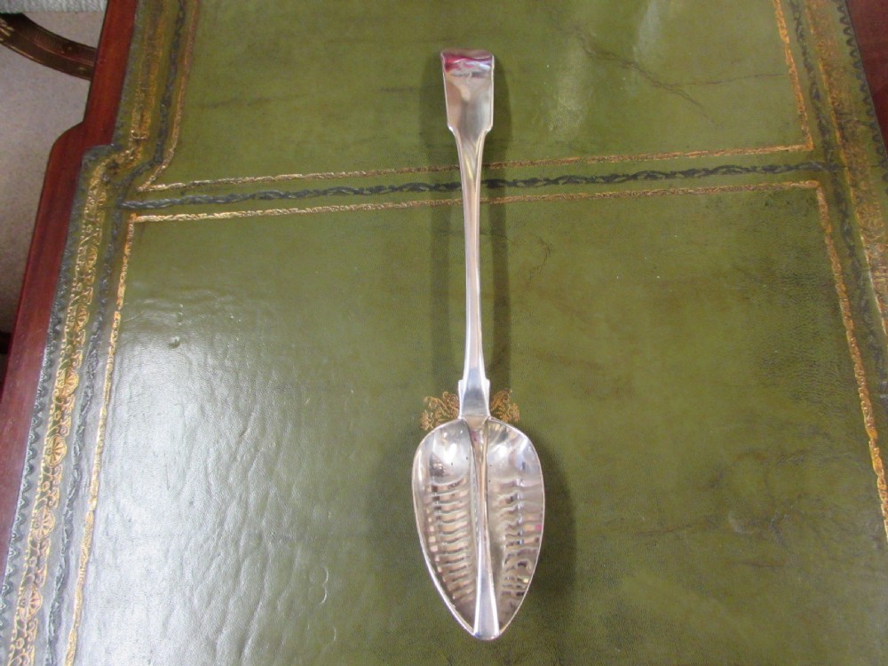A George III Irish sterling silver strainer spoon featuring a long flat handle with an engraved crest and an oval bowl with a central divider and slotted decorative piercings. View II