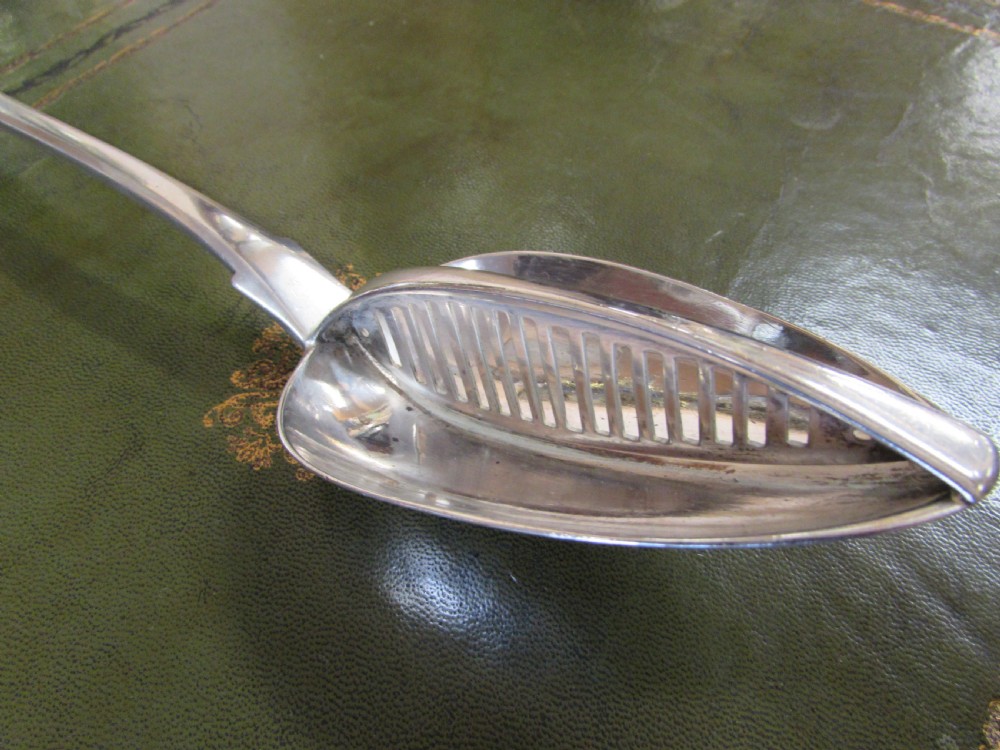 A George III Irish sterling silver strainer spoon featuring a long flat handle with an engraved crest and an oval bowl with a central divider and slotted decorative piercings. View III