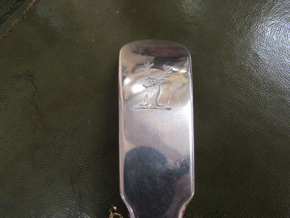 A George III Irish sterling silver strainer spoon featuring a long flat handle with an engraved crest and an oval bowl with a central divider and slotted decorative piercings. View IV