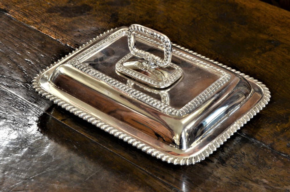 An angled close-up view of an antique rectangular silver entree dish with a matching lid and a loop handle resting on a dark wooden surface.