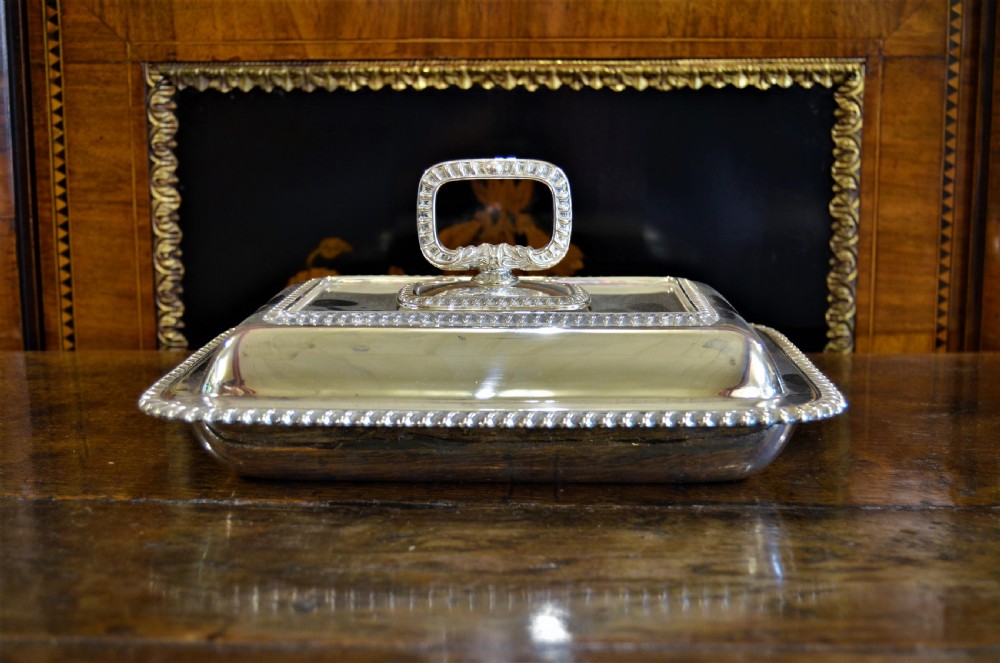 An angled close-up view of an antique rectangular silver entree dish with a matching lid and a loop handle resting on a dark wooden surface. View II