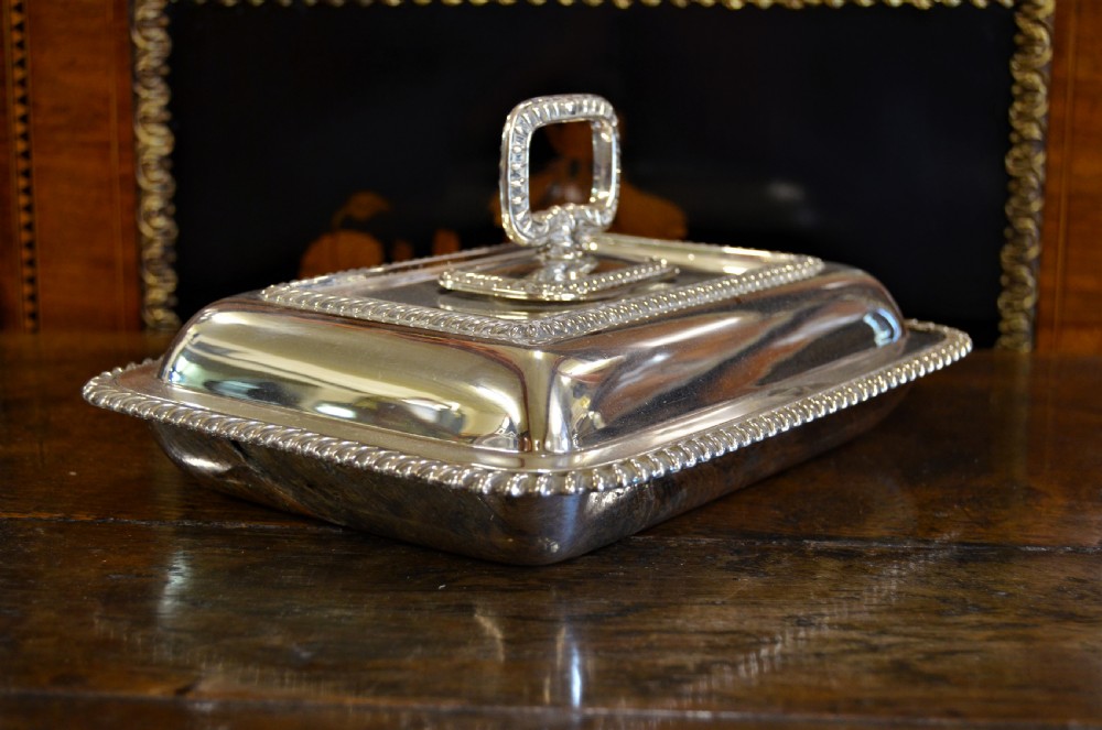 An angled close-up view of an antique rectangular silver entree dish with a matching lid and a loop handle resting on a dark wooden surface. View III
