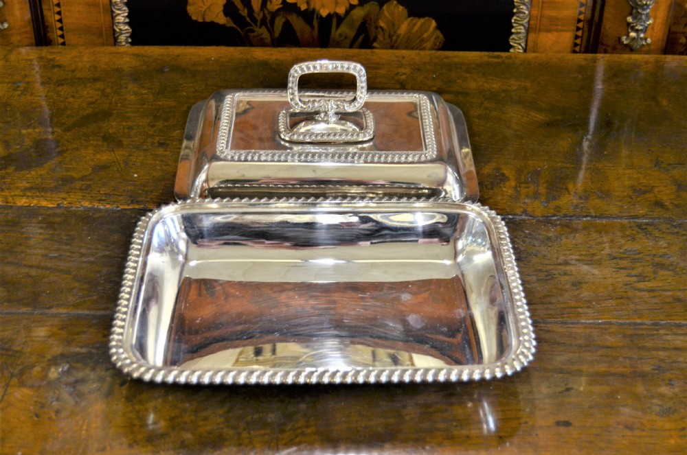 An angled close-up view of an antique rectangular silver entree dish with a matching lid and a loop handle resting on a dark wooden surface. View IV