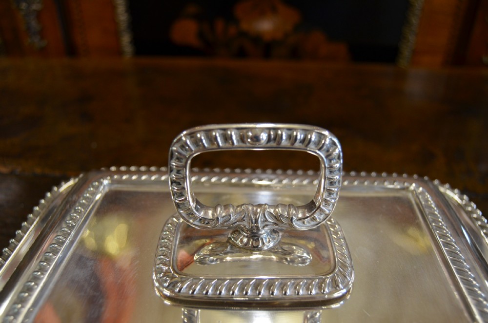 An angled close-up view of an antique rectangular silver entree dish with a matching lid and a loop handle resting on a dark wooden surface. View V