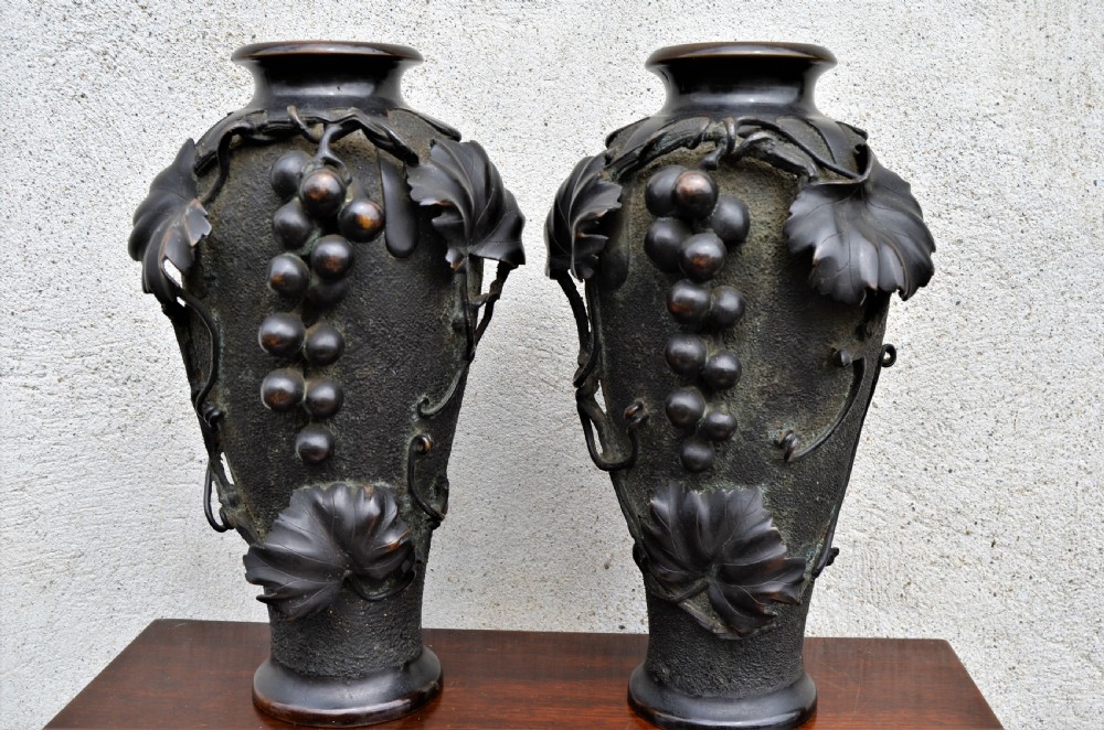 A pair of dark patinated bronze vases decorated with high-relief carved grapevine motifs and fruit clusters over a textured surface.