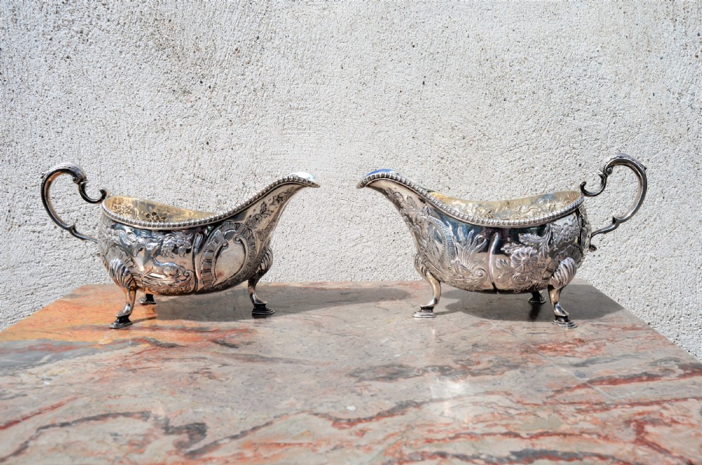 A matching pair of silver sauce boats featuring ornate floral repousse chasing, double-scroll handles, and shell-capped pad feet.