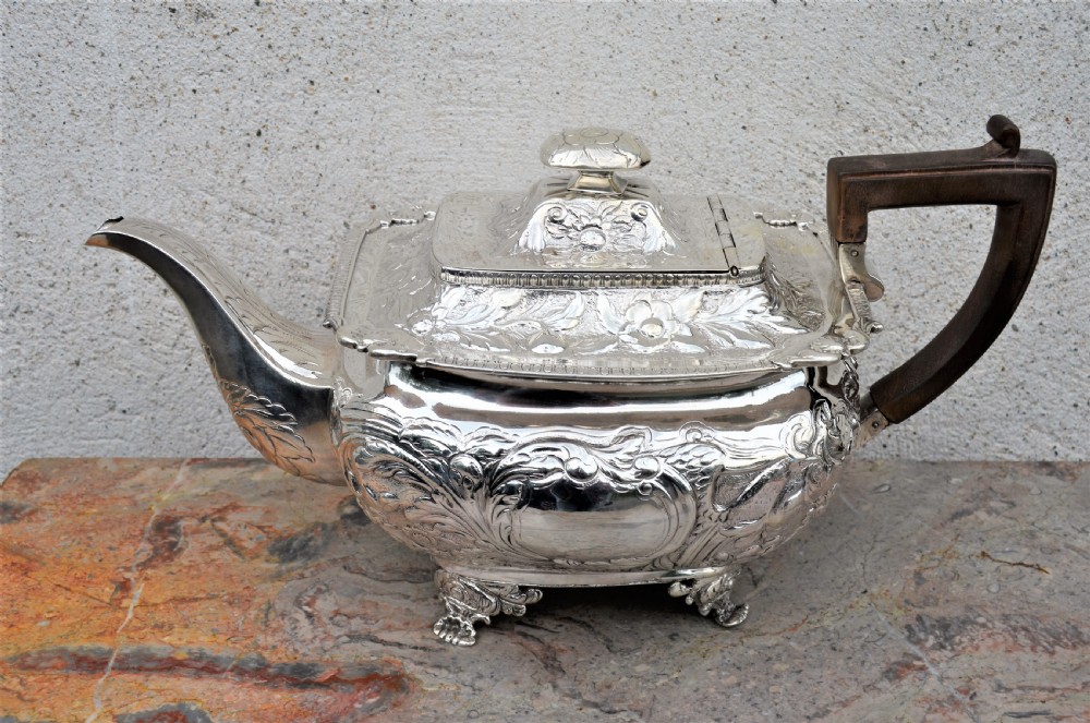 An antique sterling silver teapot featuring hand-chased floral engraving, a rectangular form resting on four feet, and a dark carved wooden handle.