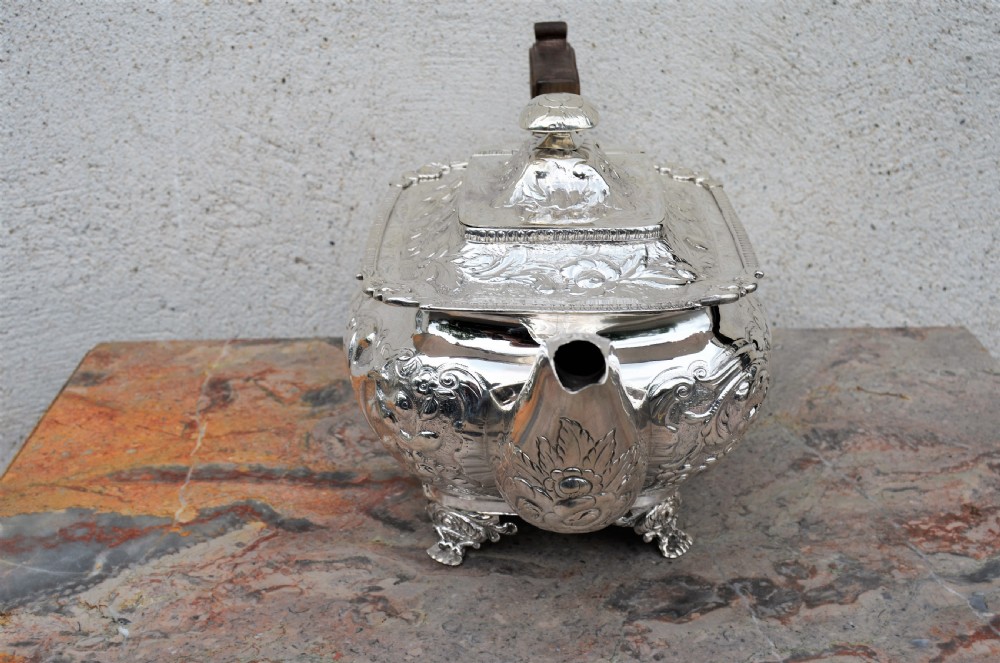 An antique sterling silver teapot featuring hand-chased floral engraving, a rectangular form resting on four feet, and a dark carved wooden handle. View II