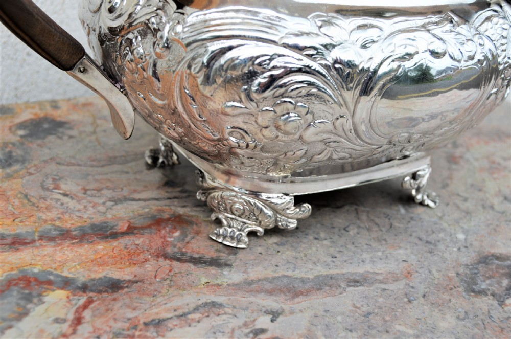 An antique sterling silver teapot featuring hand-chased floral engraving, a rectangular form resting on four feet, and a dark carved wooden handle. View IV