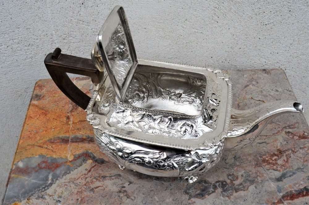An antique sterling silver teapot featuring hand-chased floral engraving, a rectangular form resting on four feet, and a dark carved wooden handle. View V