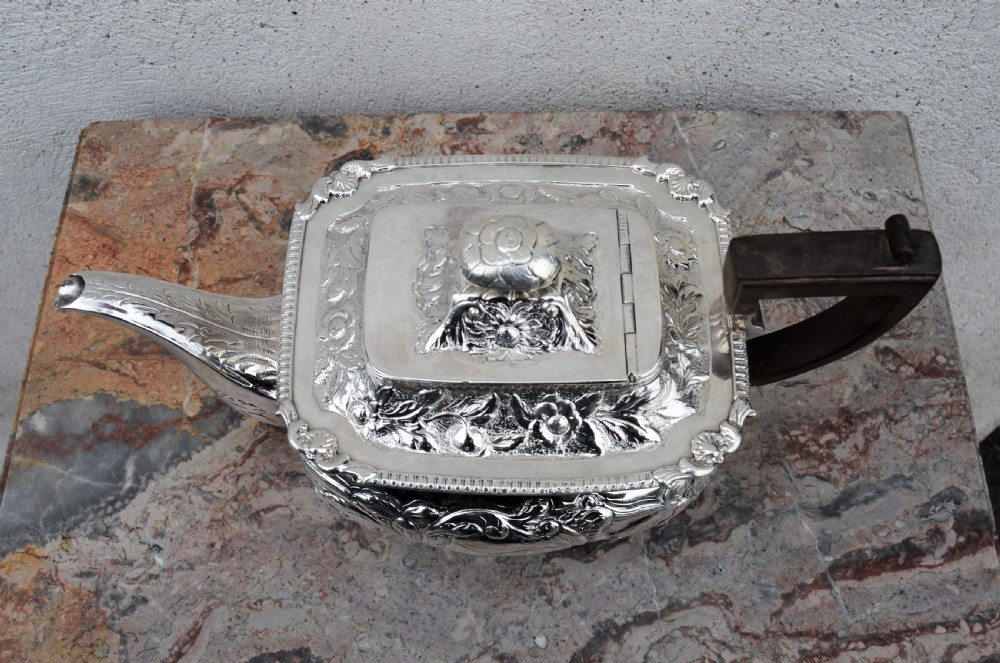 An antique sterling silver teapot featuring hand-chased floral engraving, a rectangular form resting on four feet, and a dark carved wooden handle. View VI