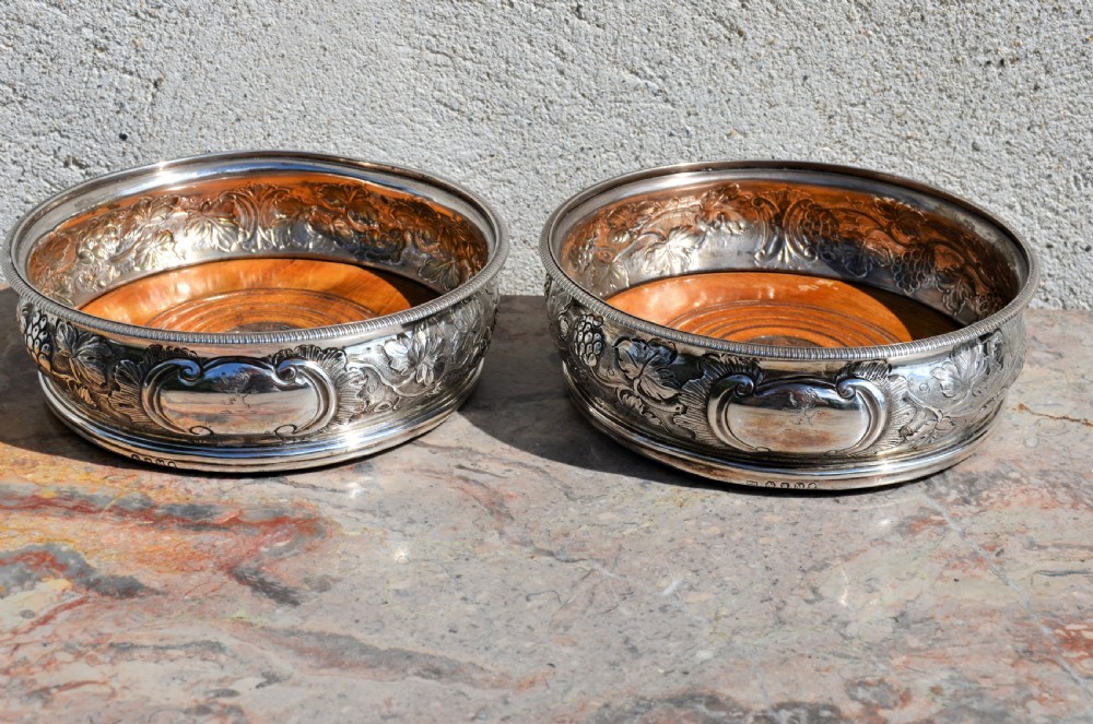 A pair of circular silver wine coasters featuring embossed botanical repousse patterns and turned wooden bases.
