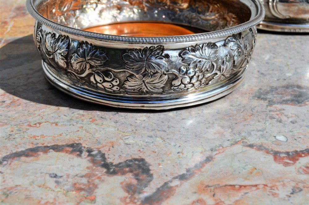 A pair of circular silver wine coasters featuring embossed botanical repousse patterns and turned wooden bases. View V