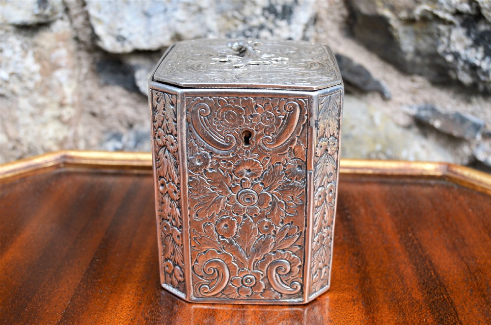 An octagonal Georgian sterling silver tea caddy decorated with intricate floral repousse patterns and featuring a keyhole on the front panel.