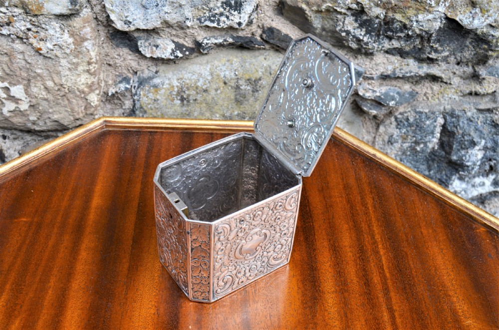 An octagonal Georgian sterling silver tea caddy decorated with intricate floral repousse patterns and featuring a keyhole on the front panel. View IV
