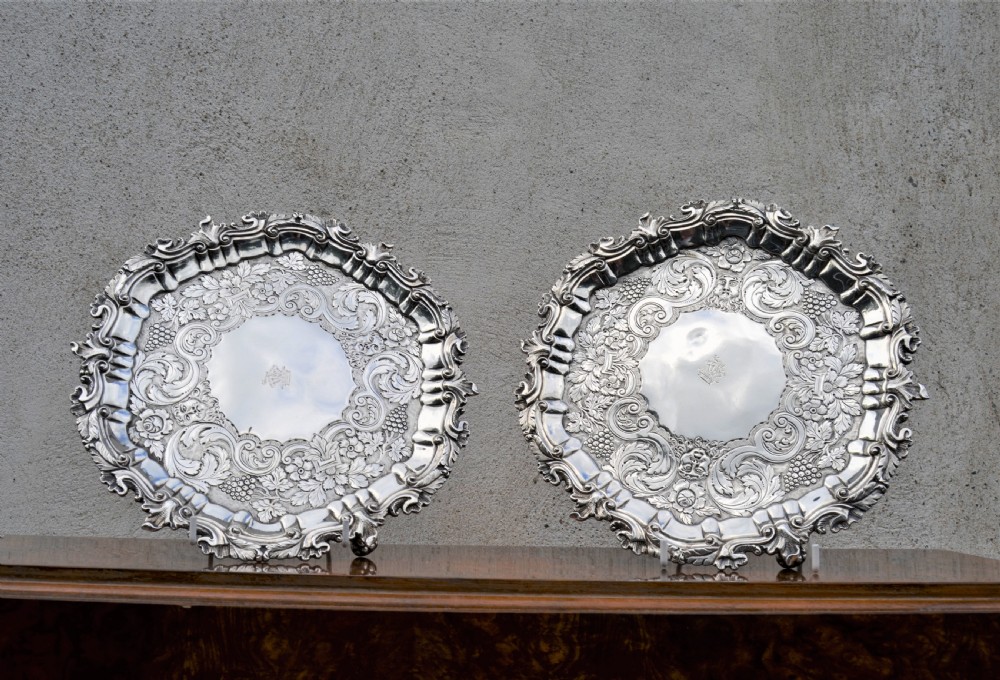 A pair of circular sterling silver salvers featuring intricate chased foliate and grapevine borders surrounding a central polished field.