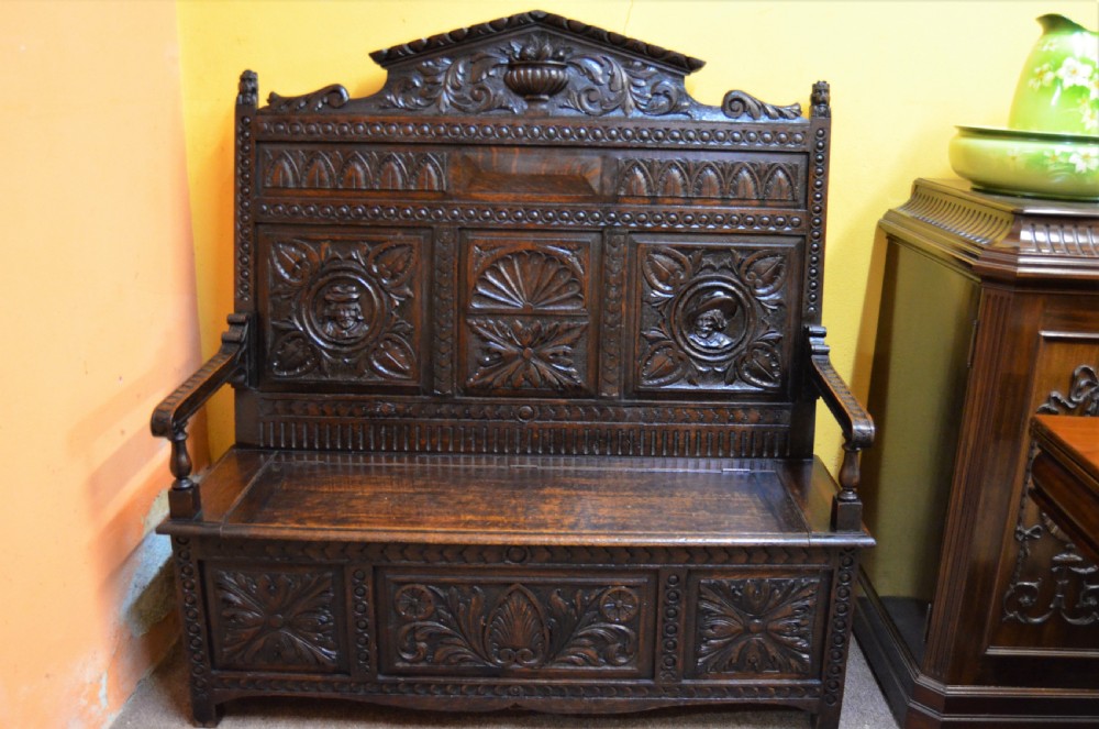 A dark Victorian carved oak monks bench with a relief-carved backrest displaying portrait medallions and foliage above a hinged box seat.