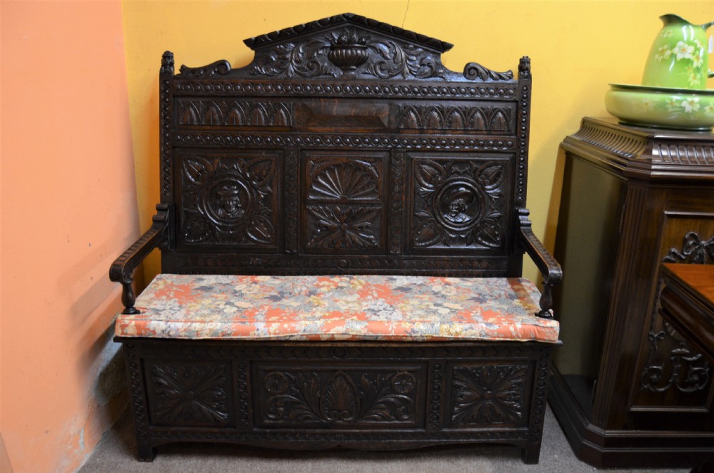 A dark Victorian carved oak monks bench with a relief-carved backrest displaying portrait medallions and foliage above a hinged box seat. View II