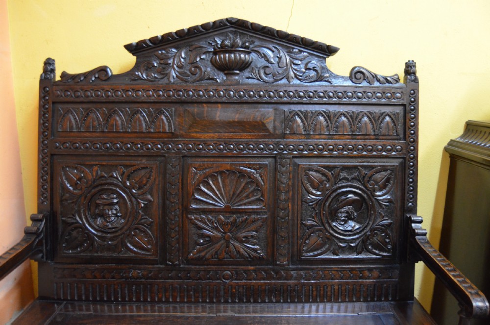 A dark Victorian carved oak monks bench with a relief-carved backrest displaying portrait medallions and foliage above a hinged box seat. View III