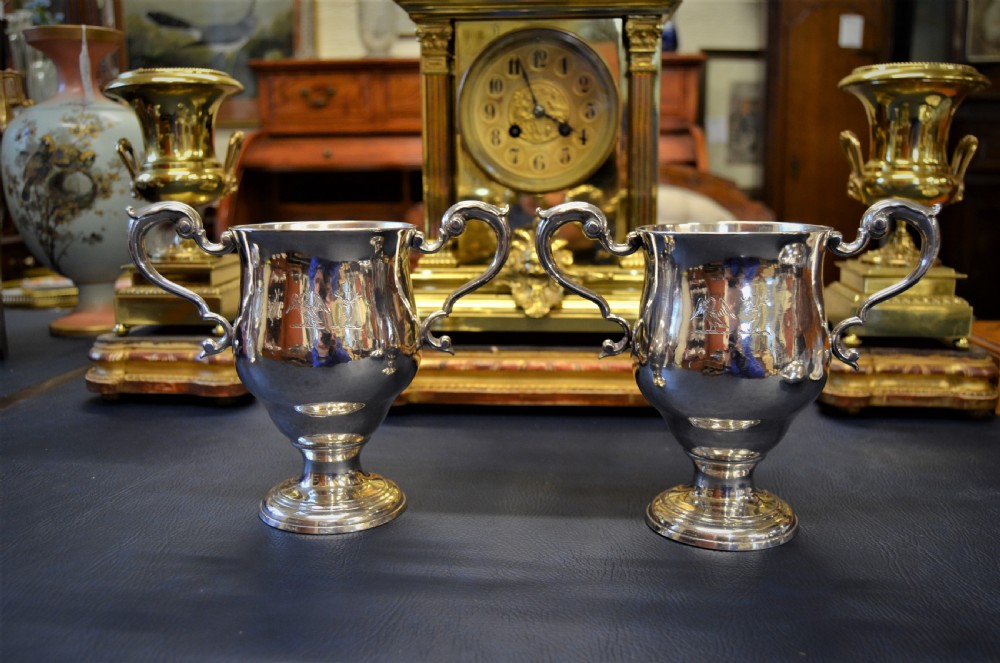 A pair of antique 18th-century silver two-handled cups with scroll handles, crest engravings, and turned pedestal bases.