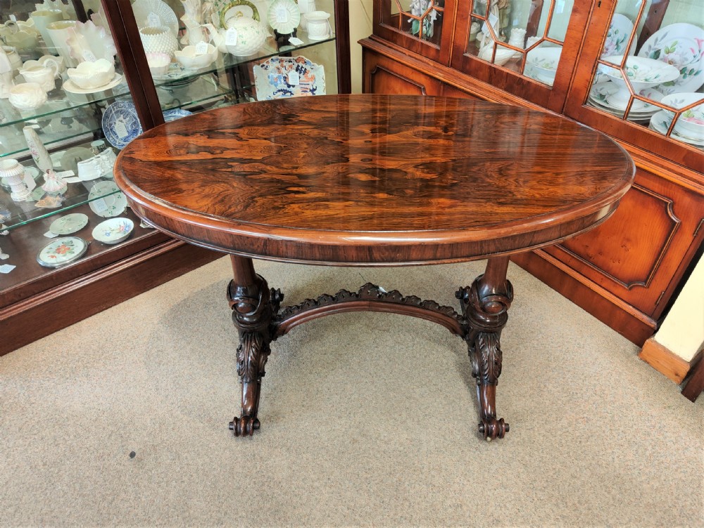 An antique Victorian oval rosewood centre table featuring a highly figured veneer top supported by carved trestle legs and a decorative stretcher.