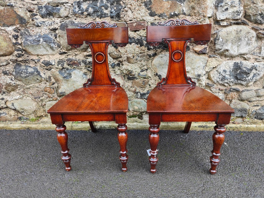 A pair of 19th-century rich brown mahogany hall chairs with carved backrests, solid seats, and turned front legs.
